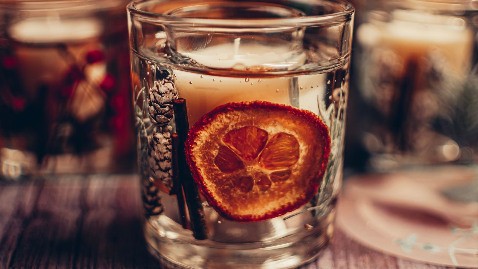 Lit candle in a glass holder with a dried orange slice and pinecone on a wooden surface.