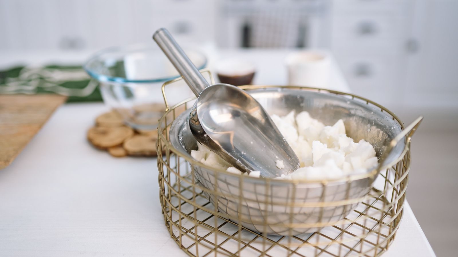 Wire basket of cream soy wax flakes with a metal scoop on a pale studio bench.