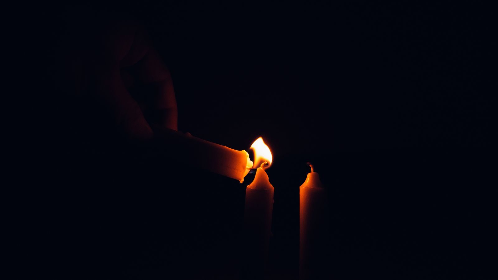 Two thin tapers being lit in low light, hands just visible behind the flames.