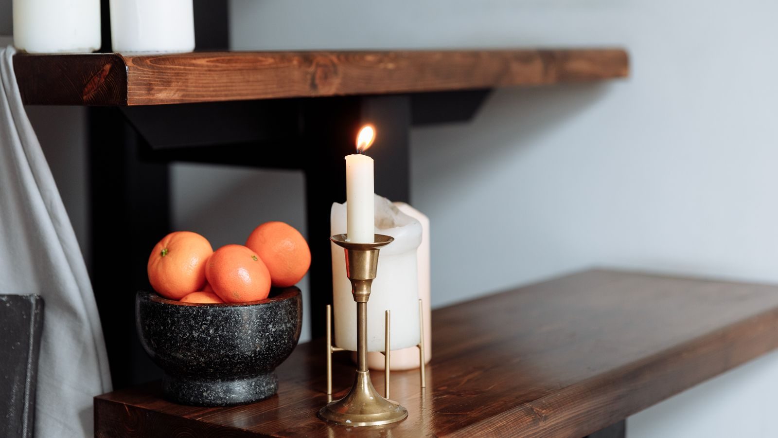 Single lit candle on a dark wood shelf with oranges in a stone bowl beside a brass candlestick.