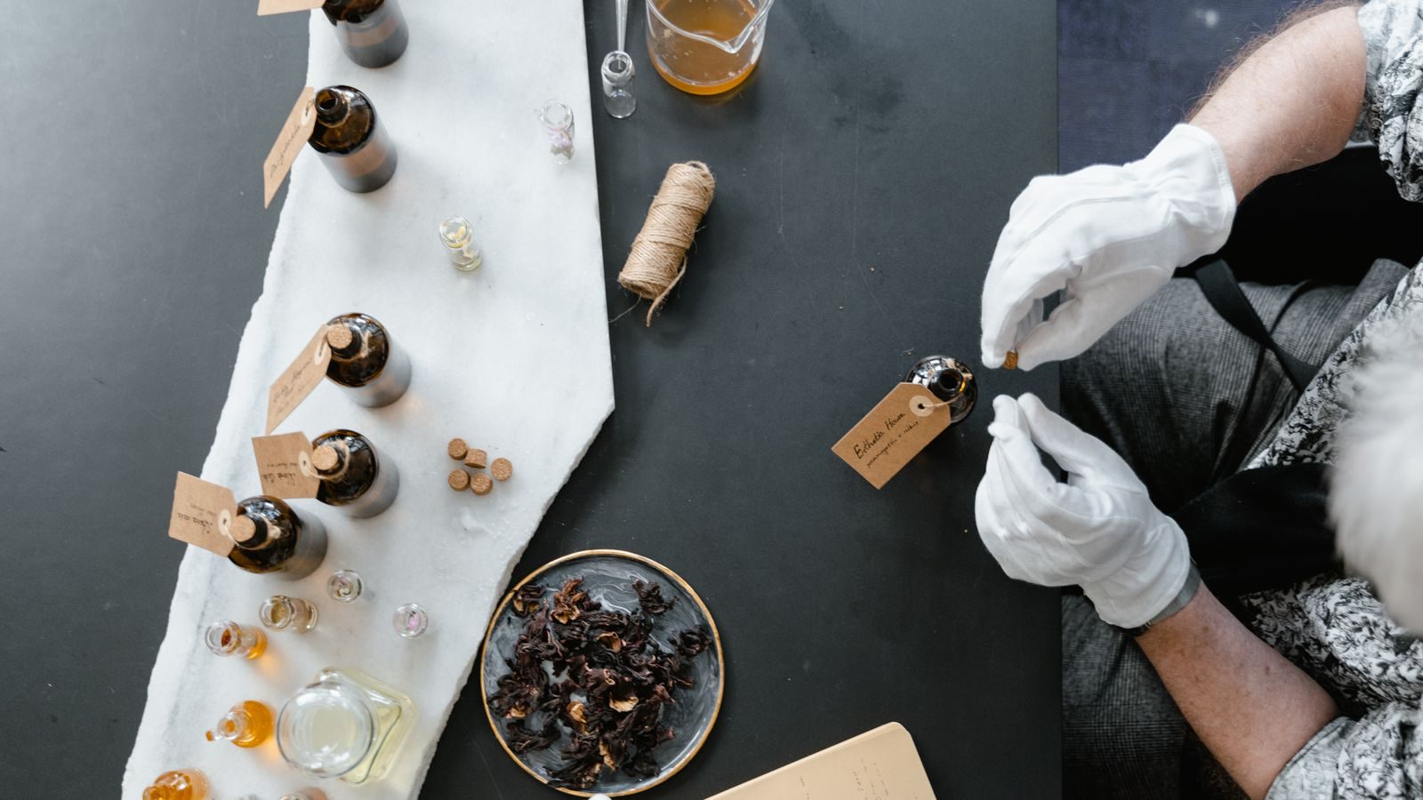 Gloved hands blending fragrance oils on a dark bench with small amber bottles and weighing dishes.