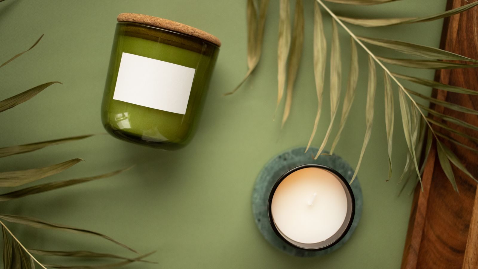 Olive-green glass jar candle with a blank kraft label, on a green surface with palm leaves.