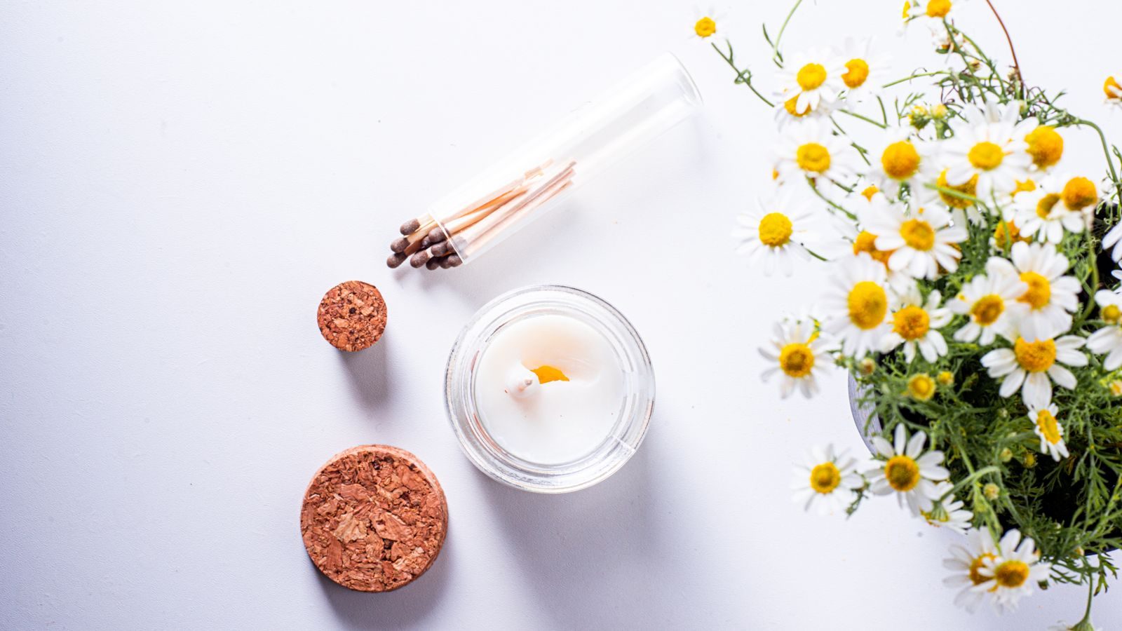 Lit tealight candle beside a small posy of chamomile flowers and matches on a white surface.